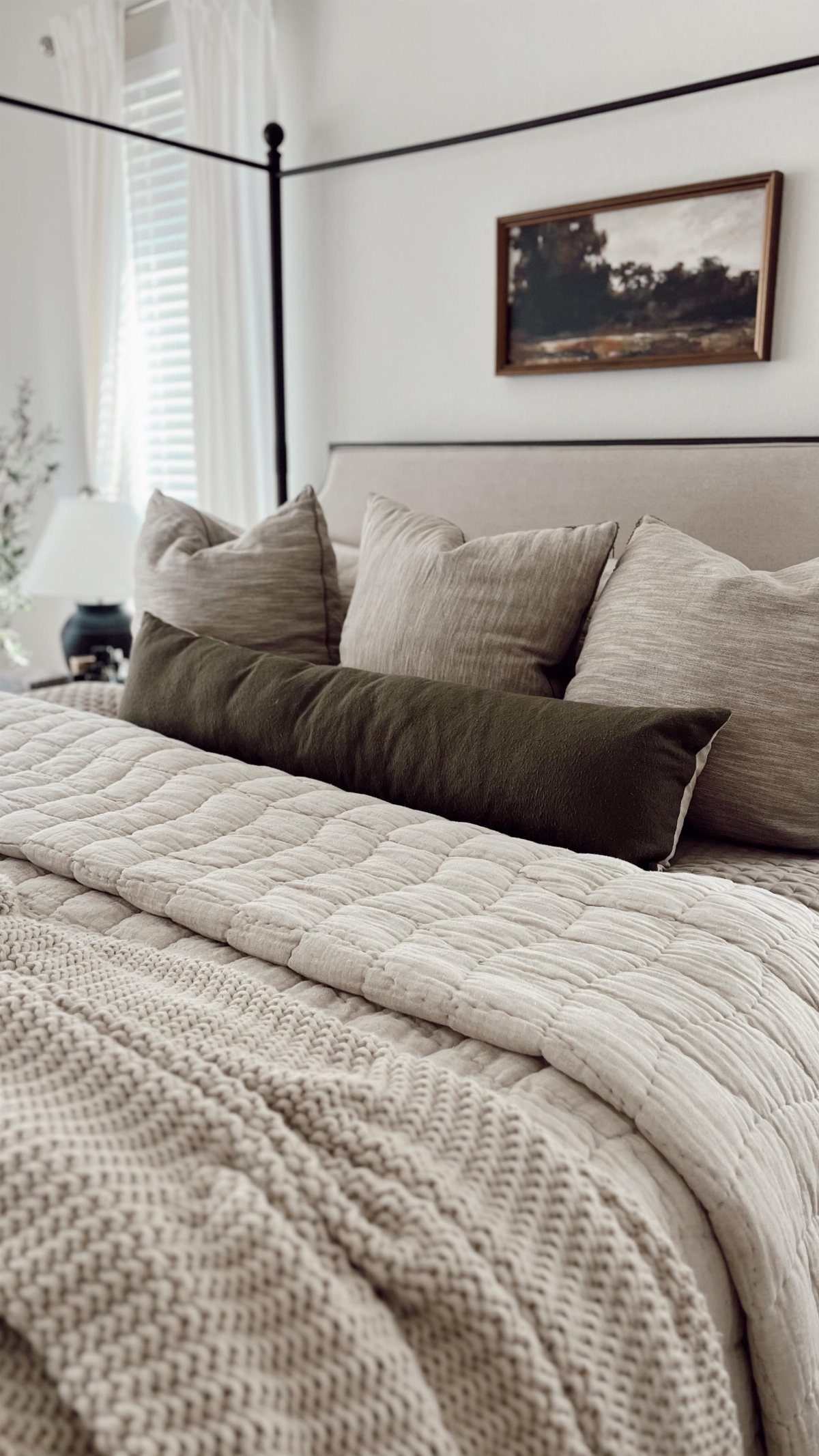 Layered neutral bedding with quilted textures, knit throws, and olive green pillows on a bed.