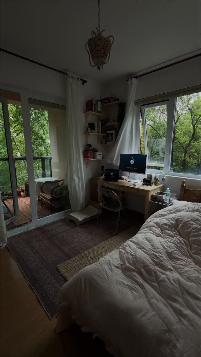 A bedroom with a desk, white bedding, and large windows overlooking green trees.
