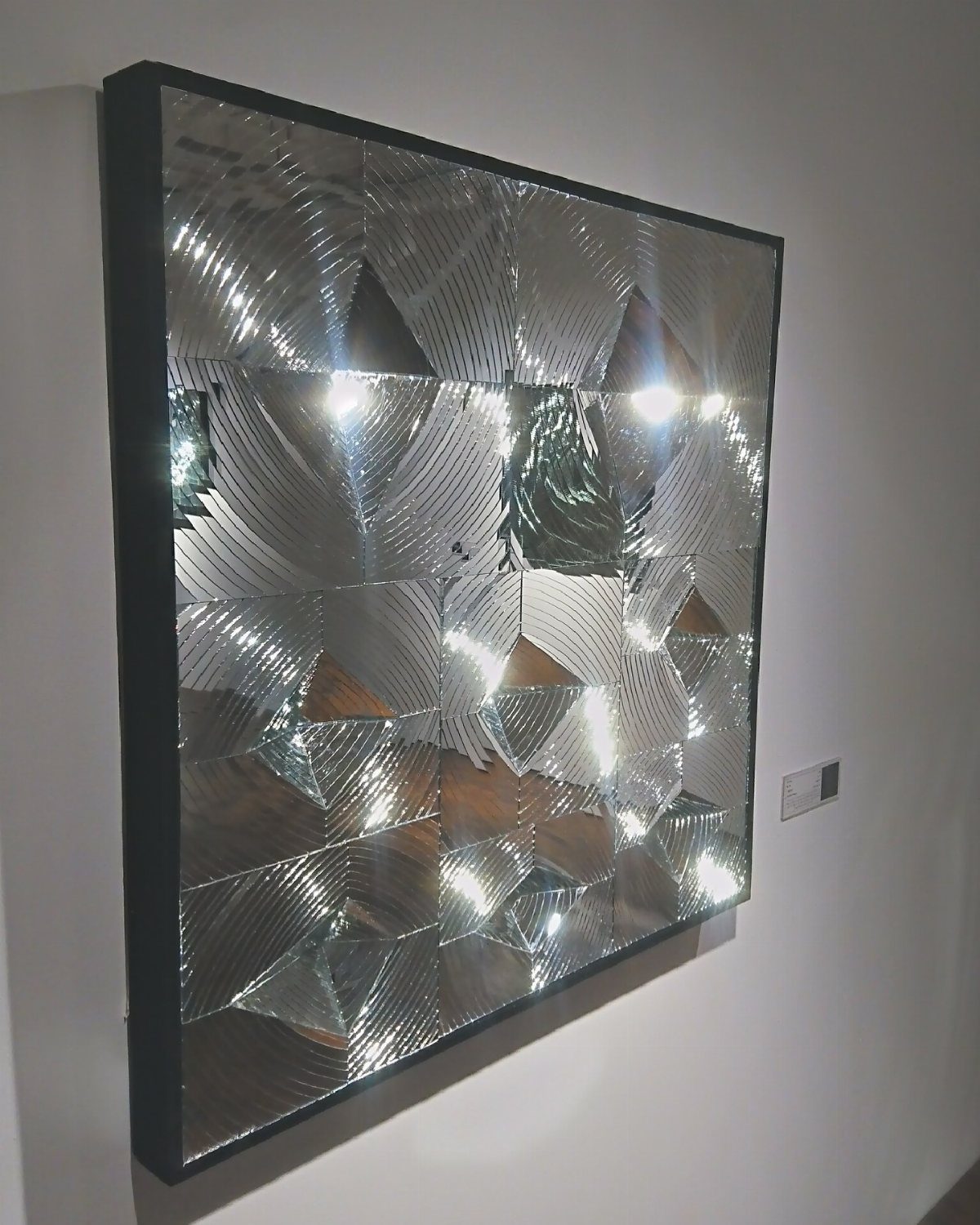 A large square black-framed mirror with faceted geometric glass panels reflecting light.