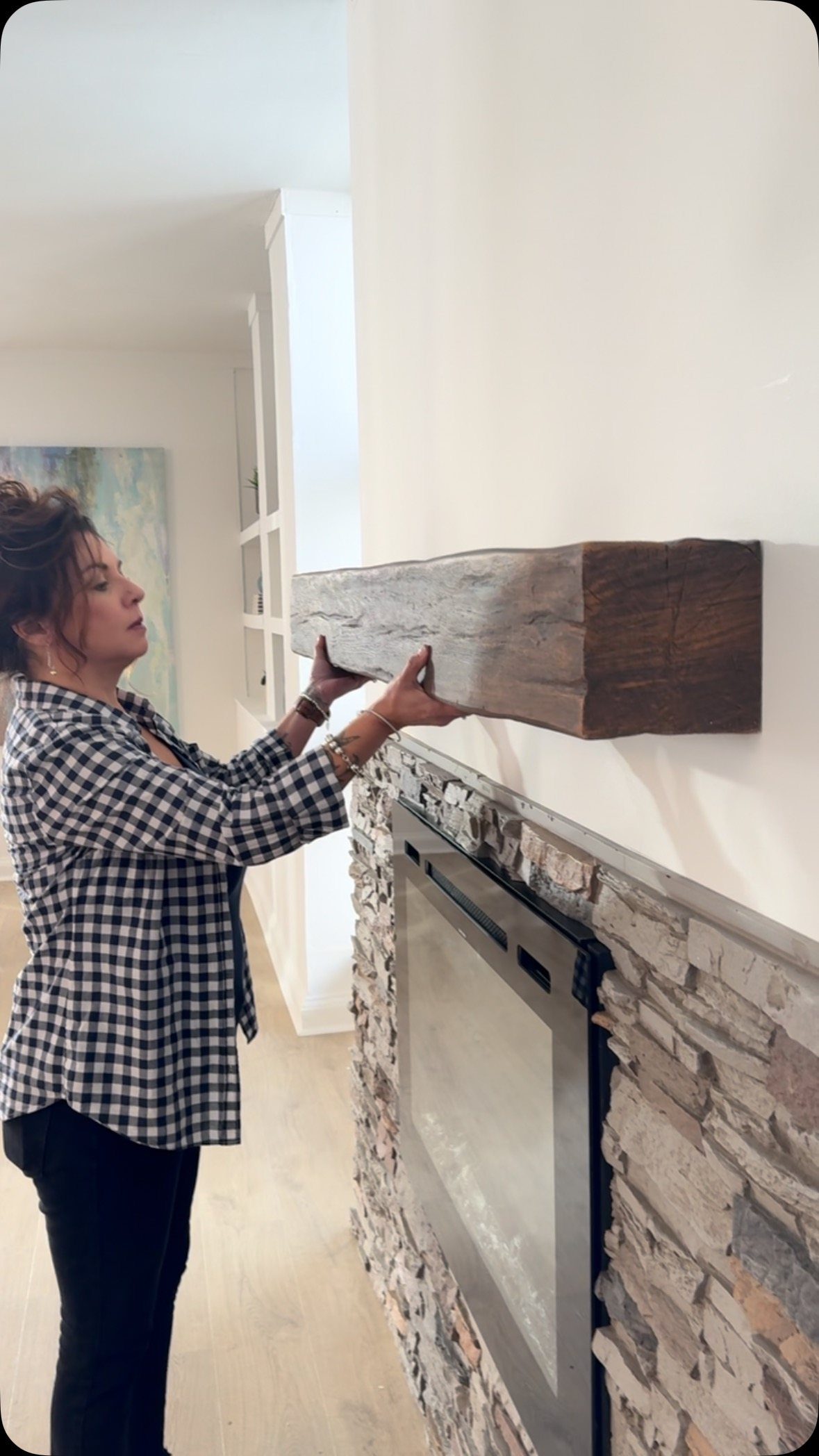 A woman installing a rustic wooden beam as a faux mantel above a stone fireplace.