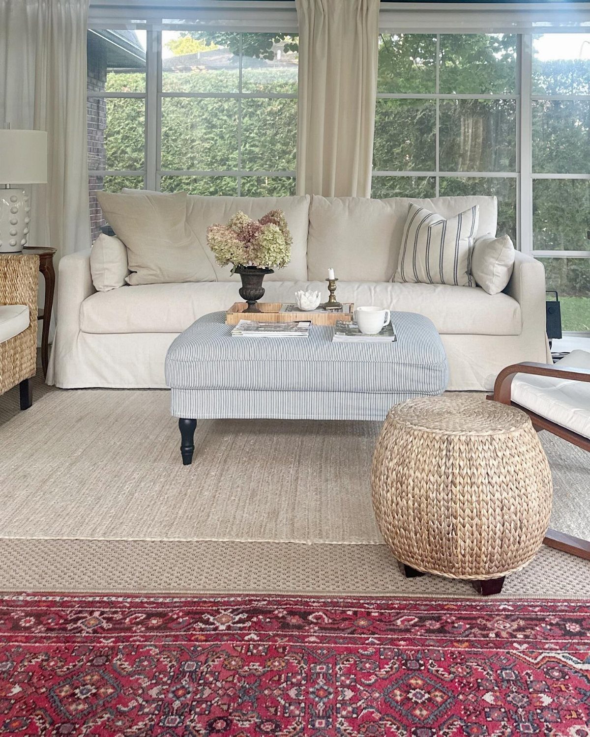 A living room showcasing layered rugs, a striped ottoman, and a woven pouf.