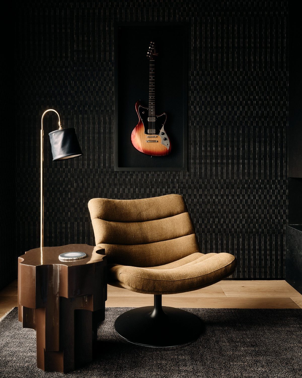 A tan lounge chair next to a geometric wooden table and a framed electric guitar on a dark wall.