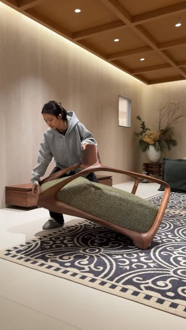 A person moving a uniquely shaped wooden lounge chair with a green cushion over a patterned rug.