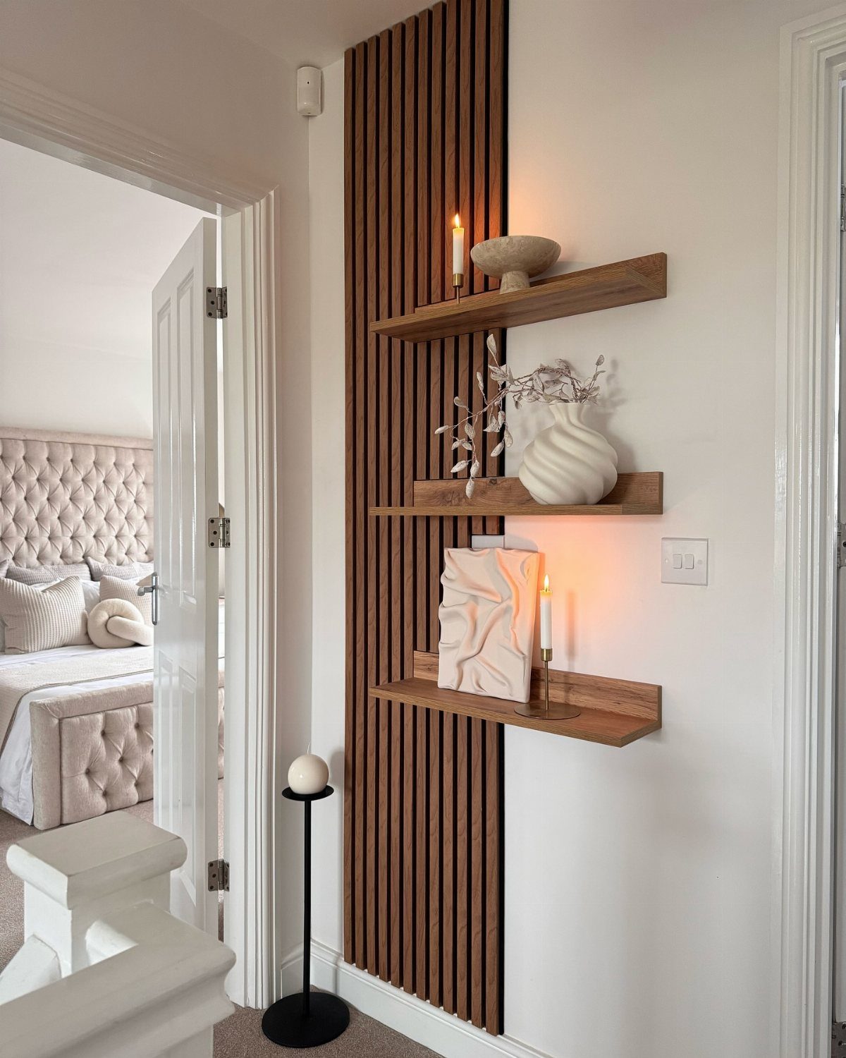 Wooden slat wall with floating shelves holding white ceramics and lit candles in a hallway.