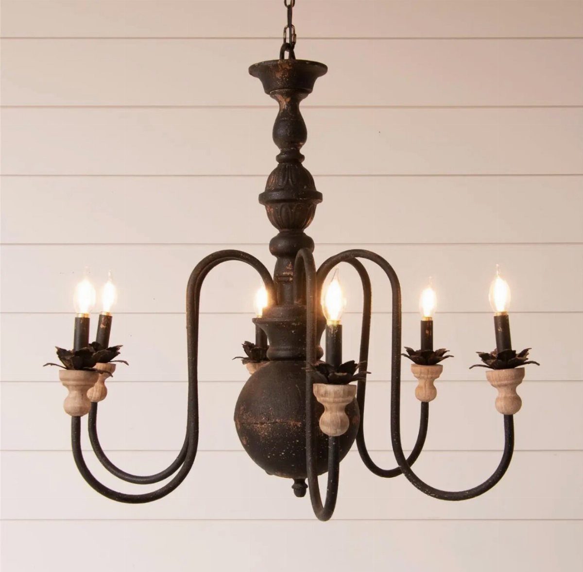 A rustic dark metal chandelier with candle lights against a white shiplap wall.