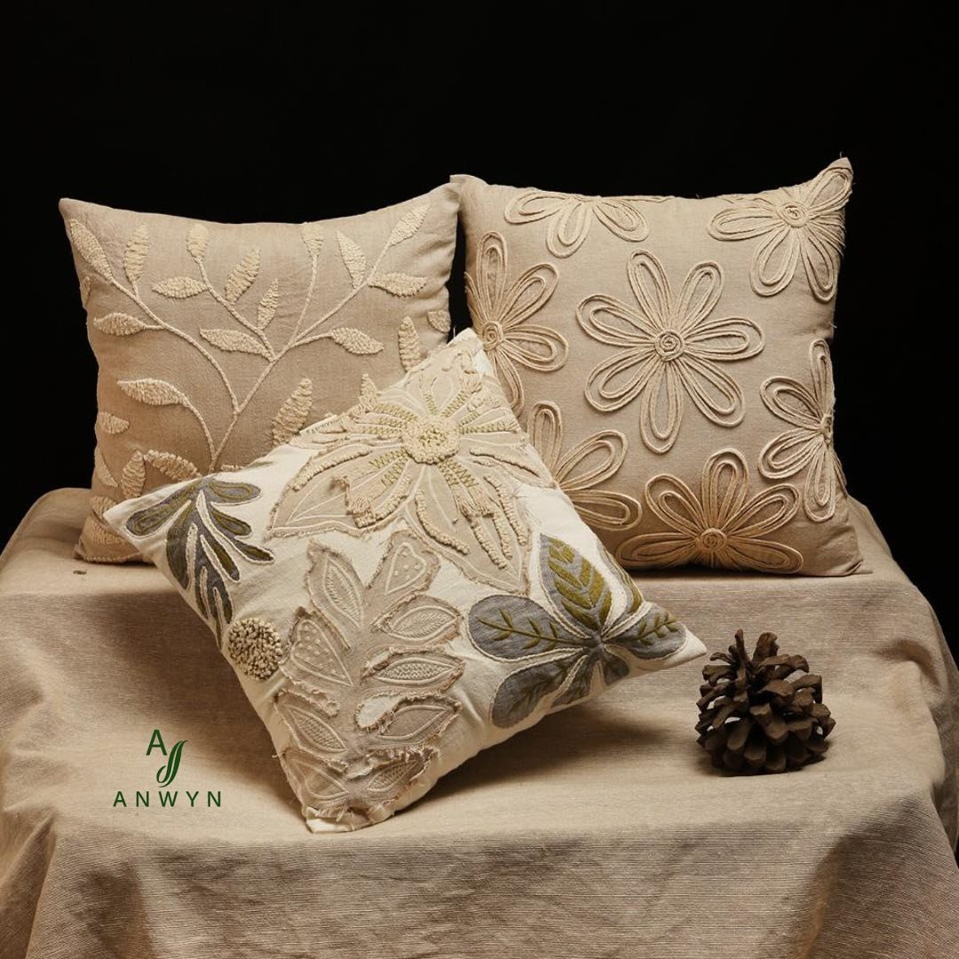 Three embroidered floral pillows in neutral tones resting on a linen cloth with a pinecone.