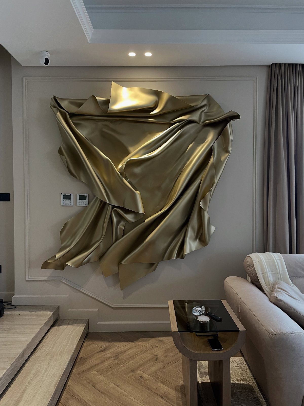 Large gold metallic sculptural art piece mounted on a grey paneled living room wall.
