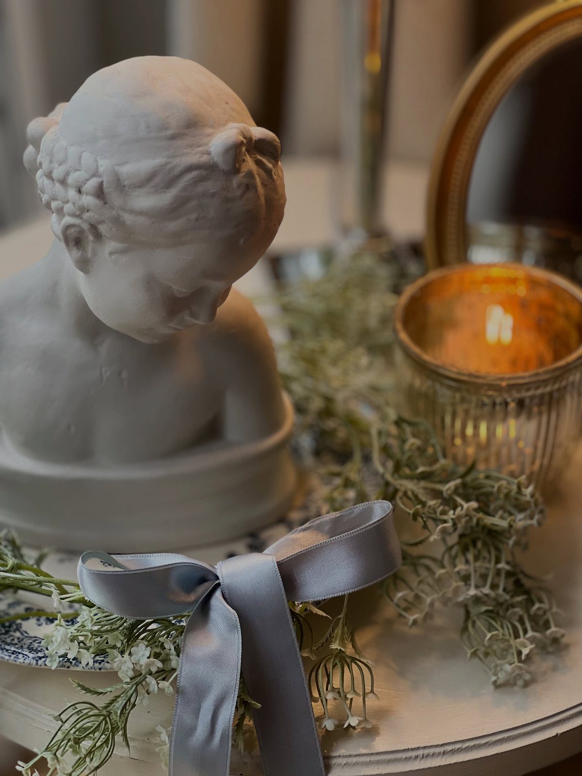 White classical bust sculpture with a grey satin ribbon and greenery on a table.