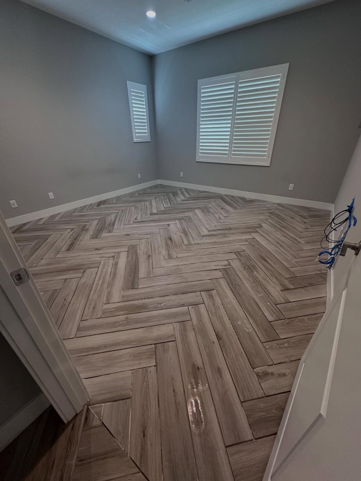 Herringbone pattern wood flooring in an empty room with grey walls and white shutters.