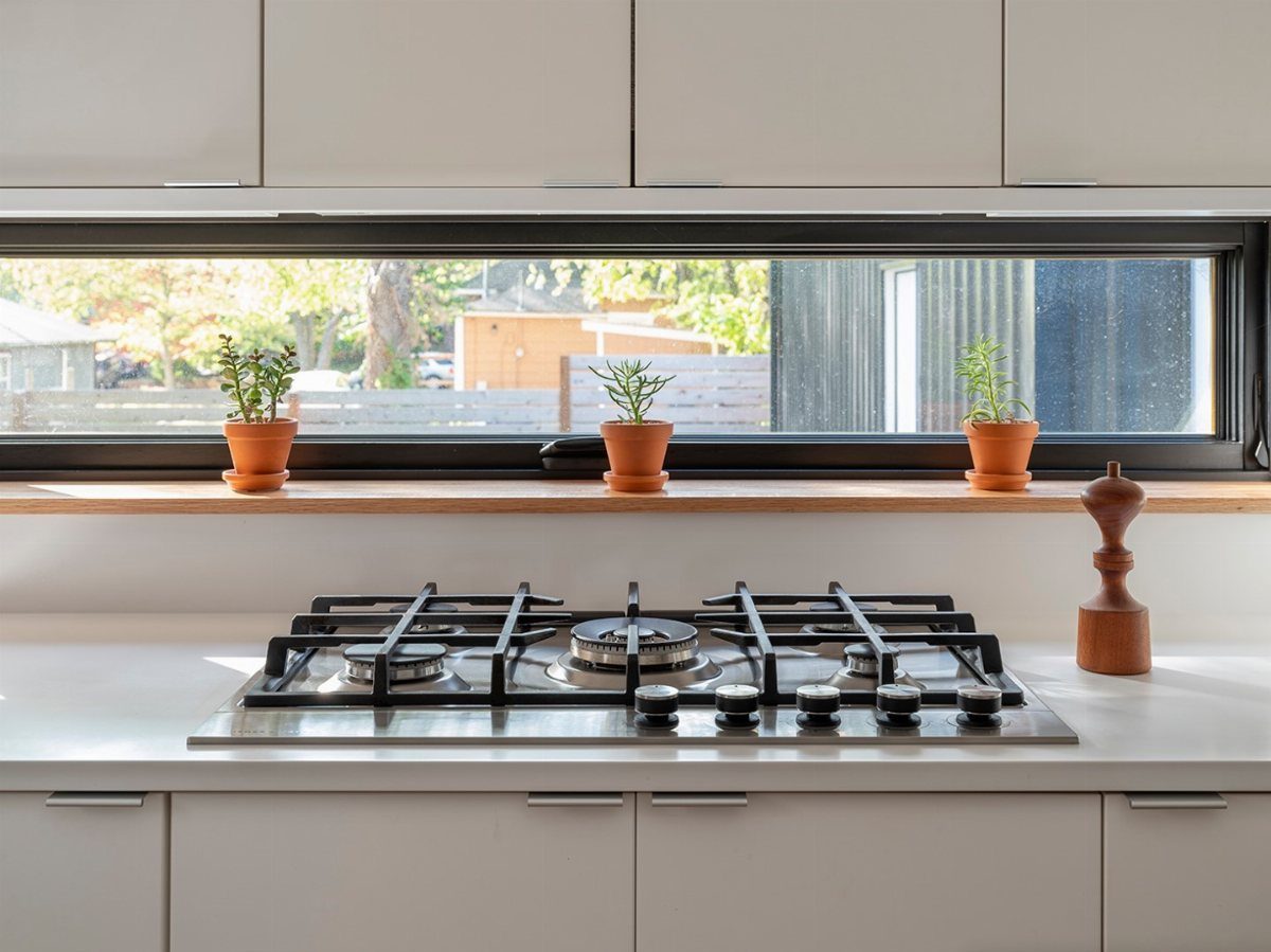 A modern white kitchen with a long horizontal window acting as a backsplash above a stovetop.
