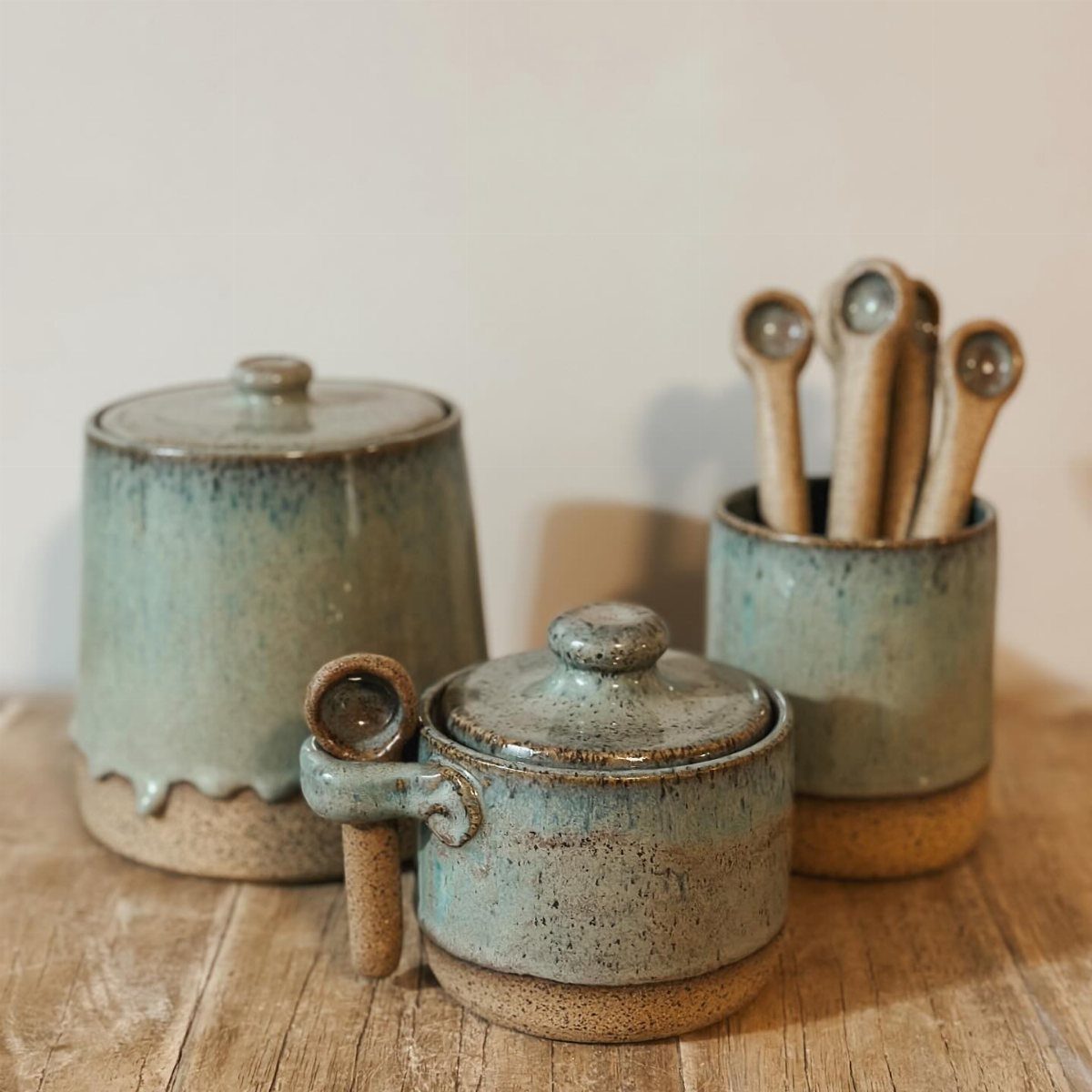 Handmade blue-green ceramic jars and spoons resting on a rustic wooden surface.