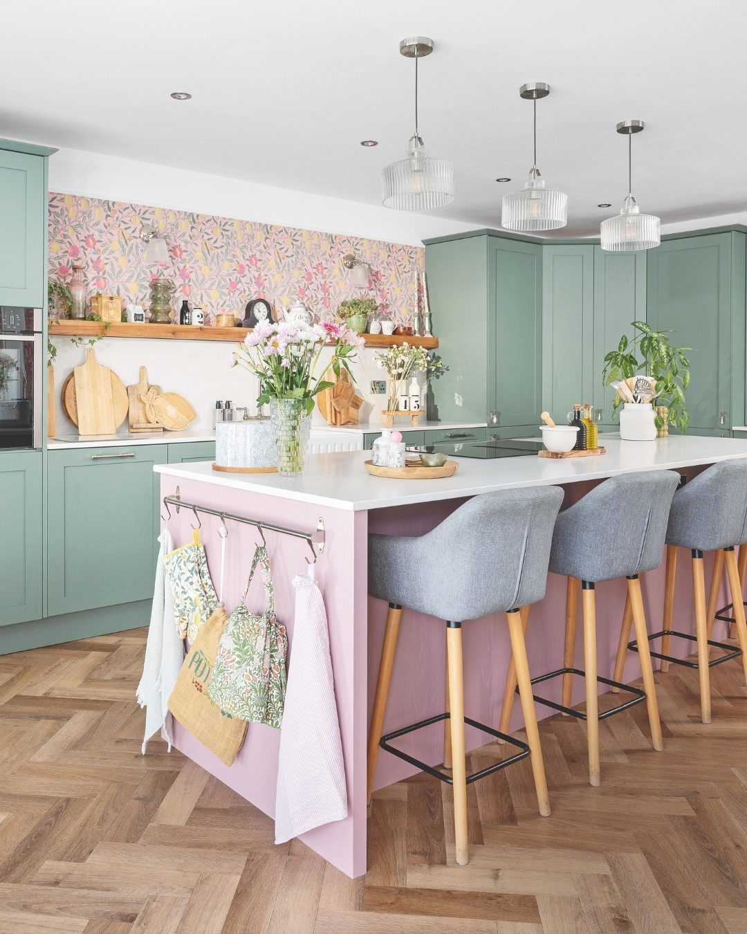 A bright kitchen with sage green cabinets, a pink island, and bold floral wallpaper.