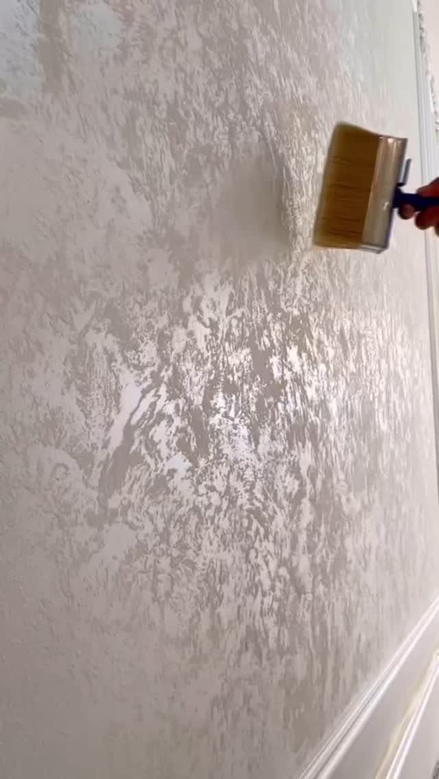 A hand applying textured silver paint to a wall with a wide brush.