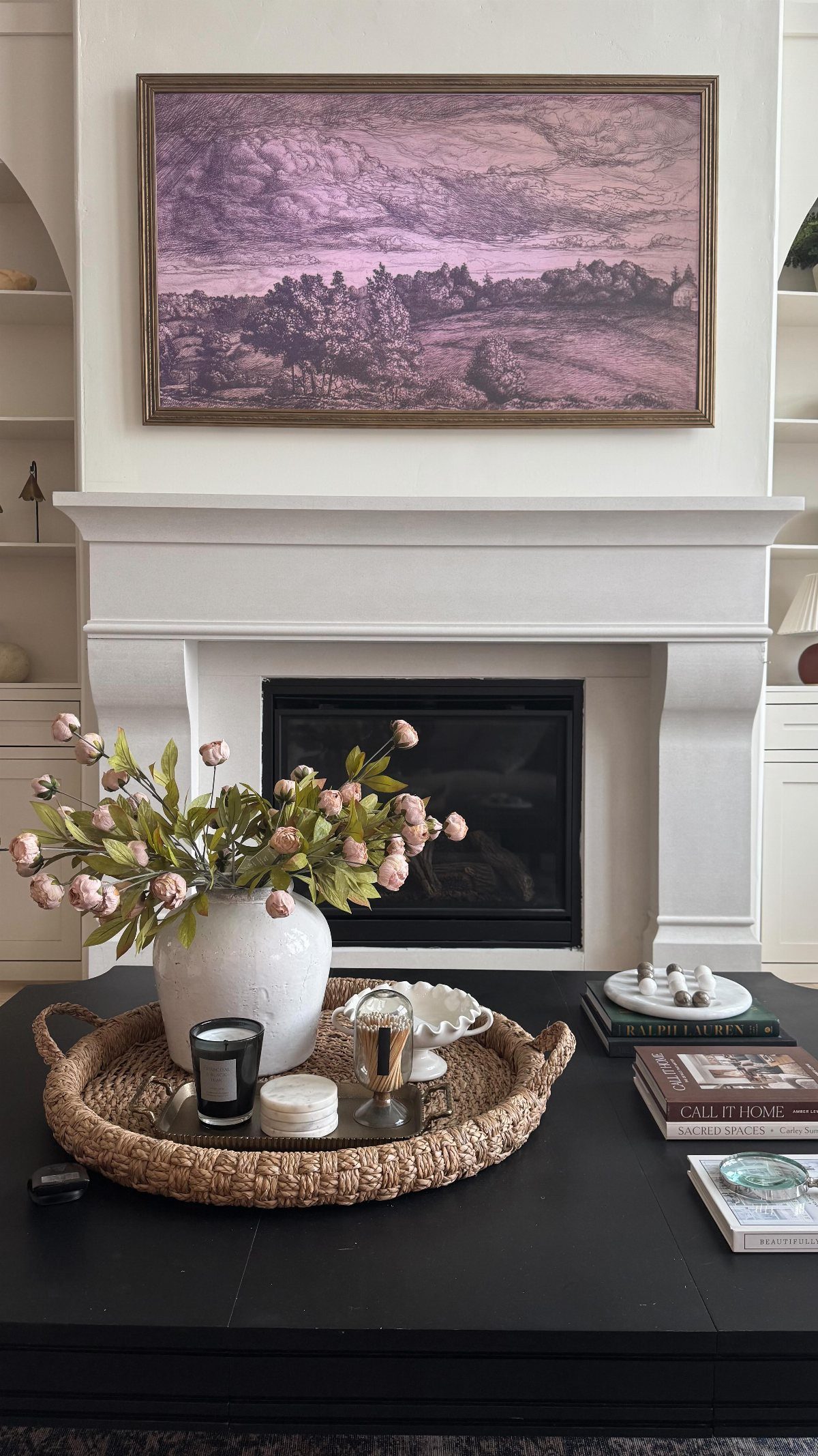 White fireplace mantel with a large landscape painting and a coffee table with floral decor.
