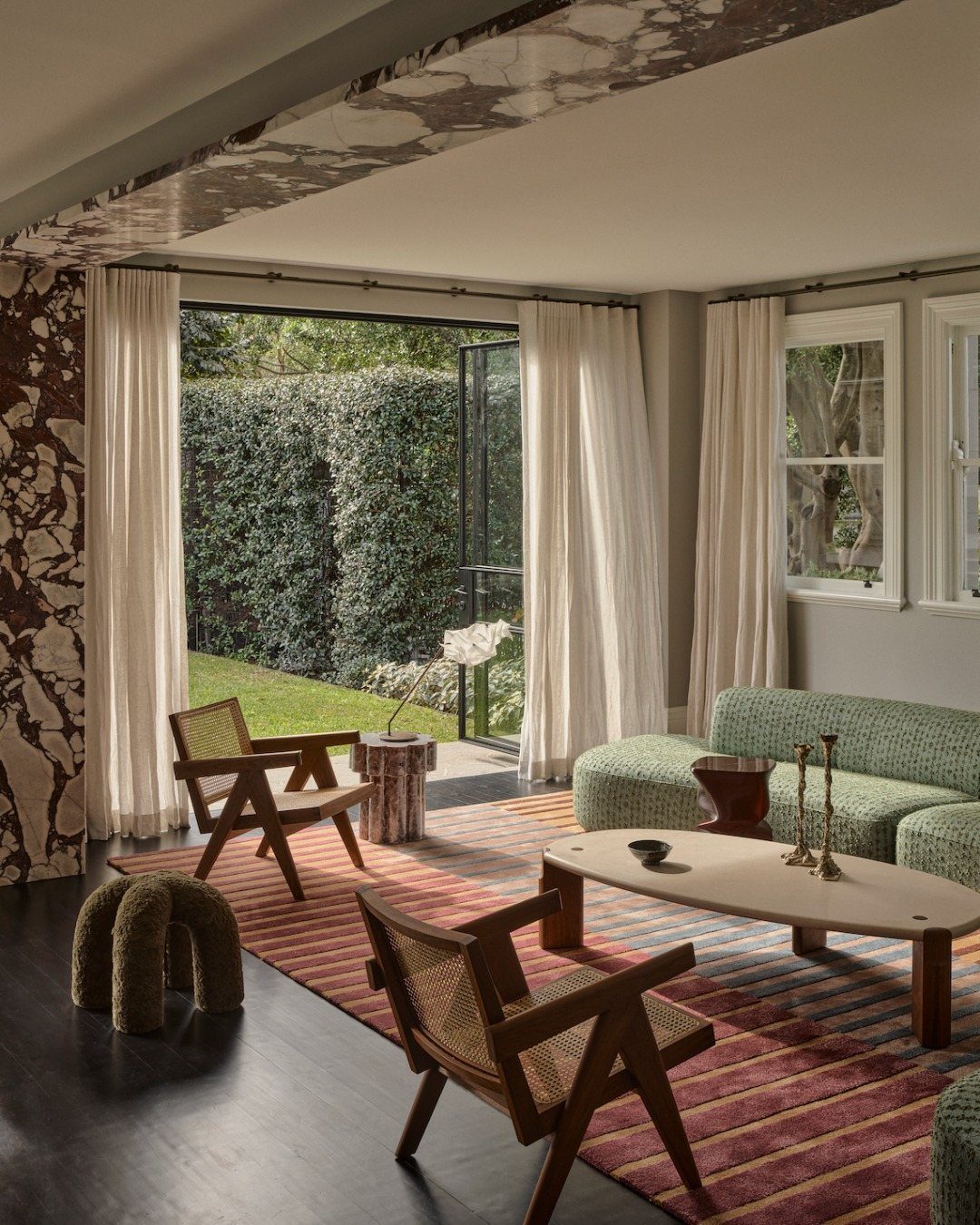 Modern living room with green textured sofa, wooden chairs, and a striped rug overlooking a garden.