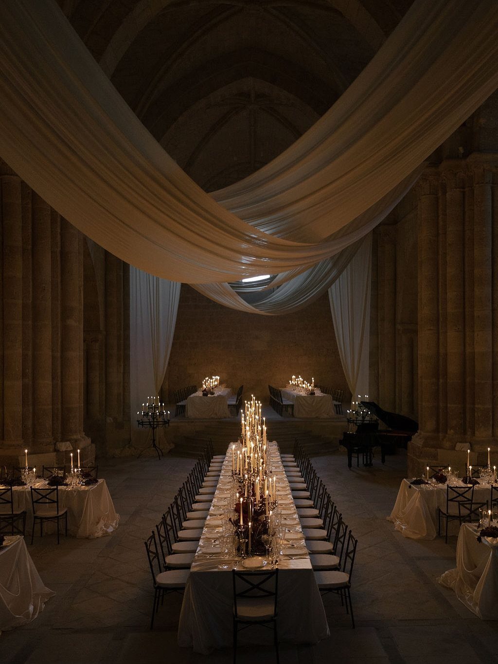 Long dining table lined with many glowing taper candles in a grand stone hall.