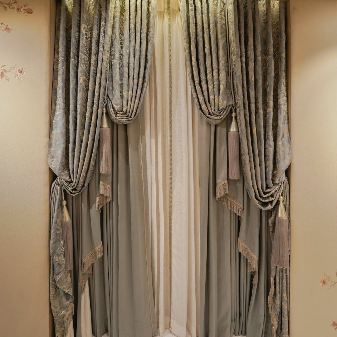 Ornate grey patterned drapery with decorative tassels hanging over a window with sheer curtains.