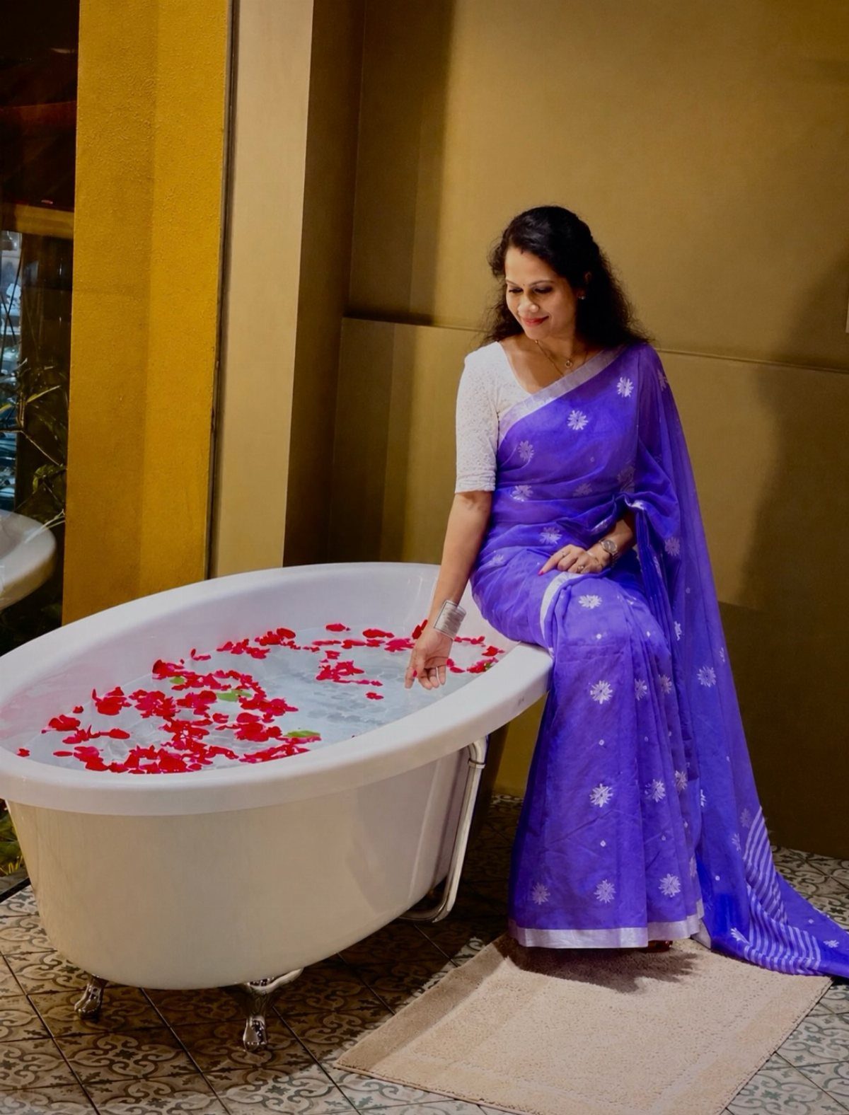 A woman in a purple sari sitting by a white bathtub filled with red flower petals.