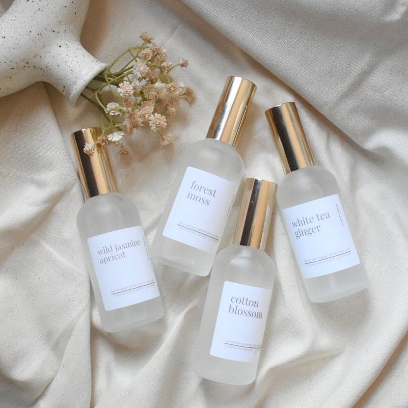 Four frosted glass scent spray bottles with gold caps resting on cream fabric.