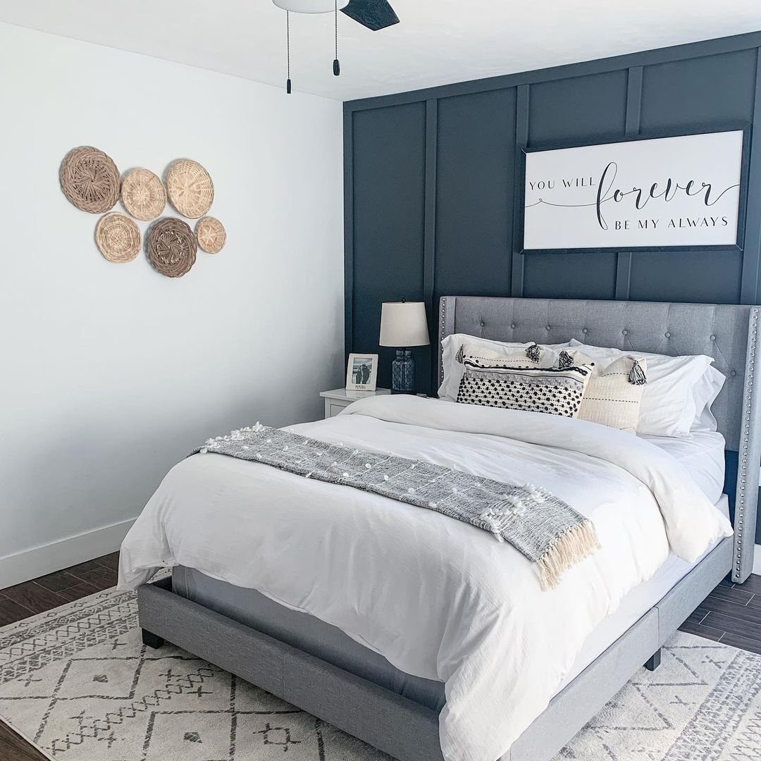 A grey tufted bed with white linens positioned against a navy blue paneled accent wall.