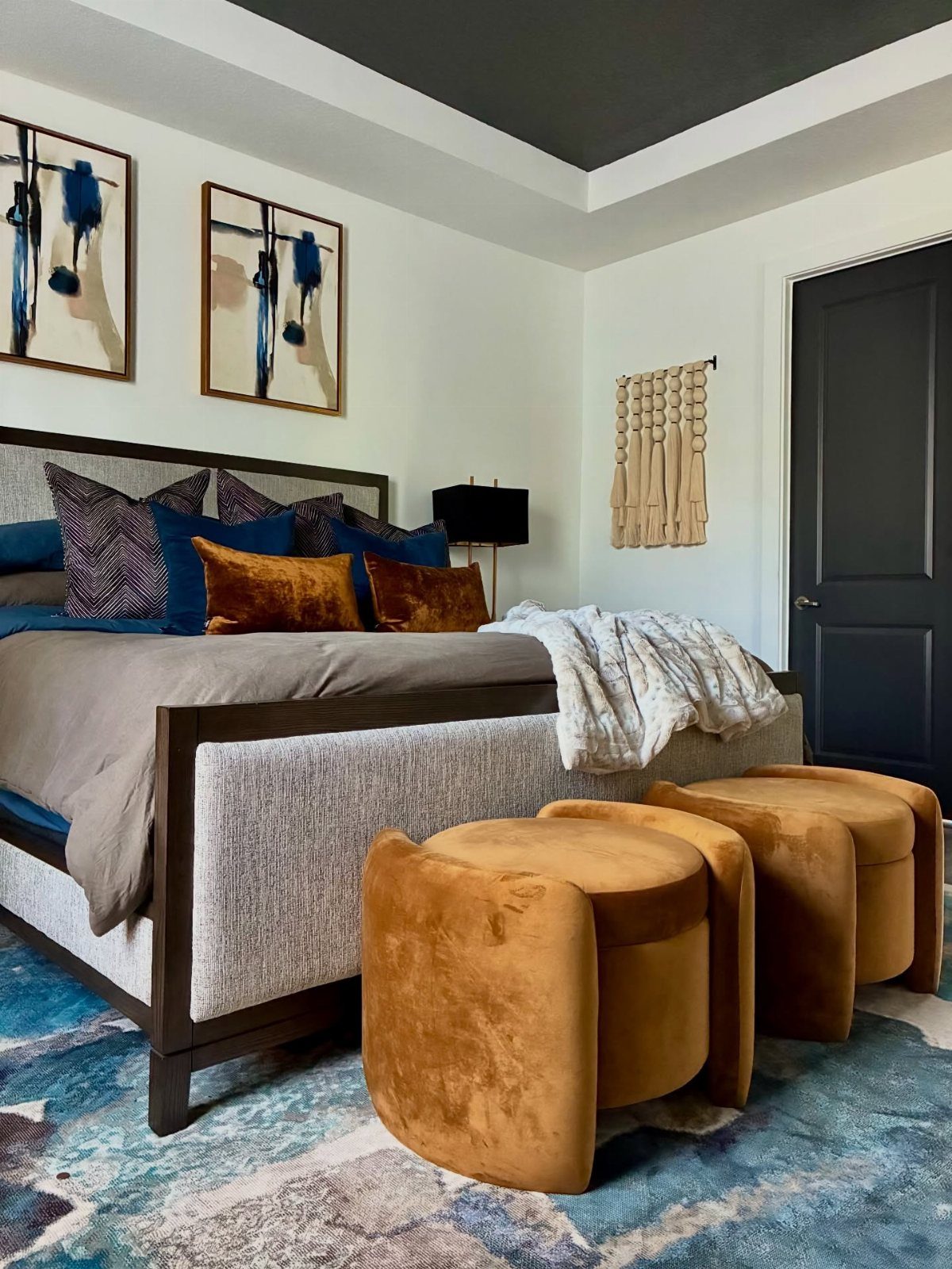 A modern bedroom with a dark wood bed frame, ochre velvet ottomans, and blue patterned rug.