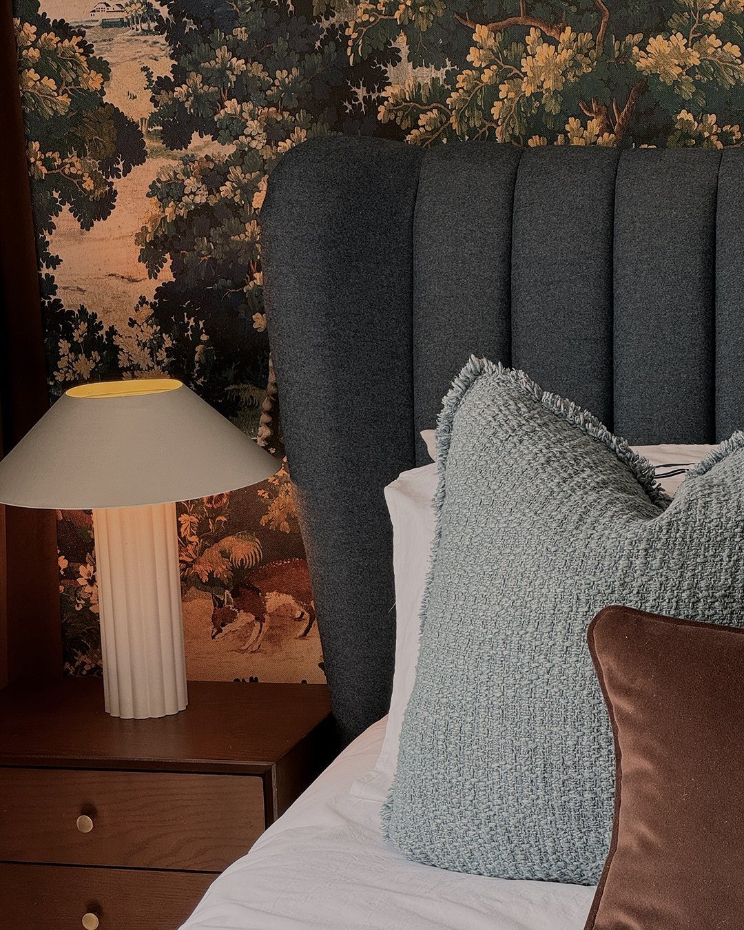 Navy blue tufted headboard against landscape wallpaper with a wooden nightstand and lamp.