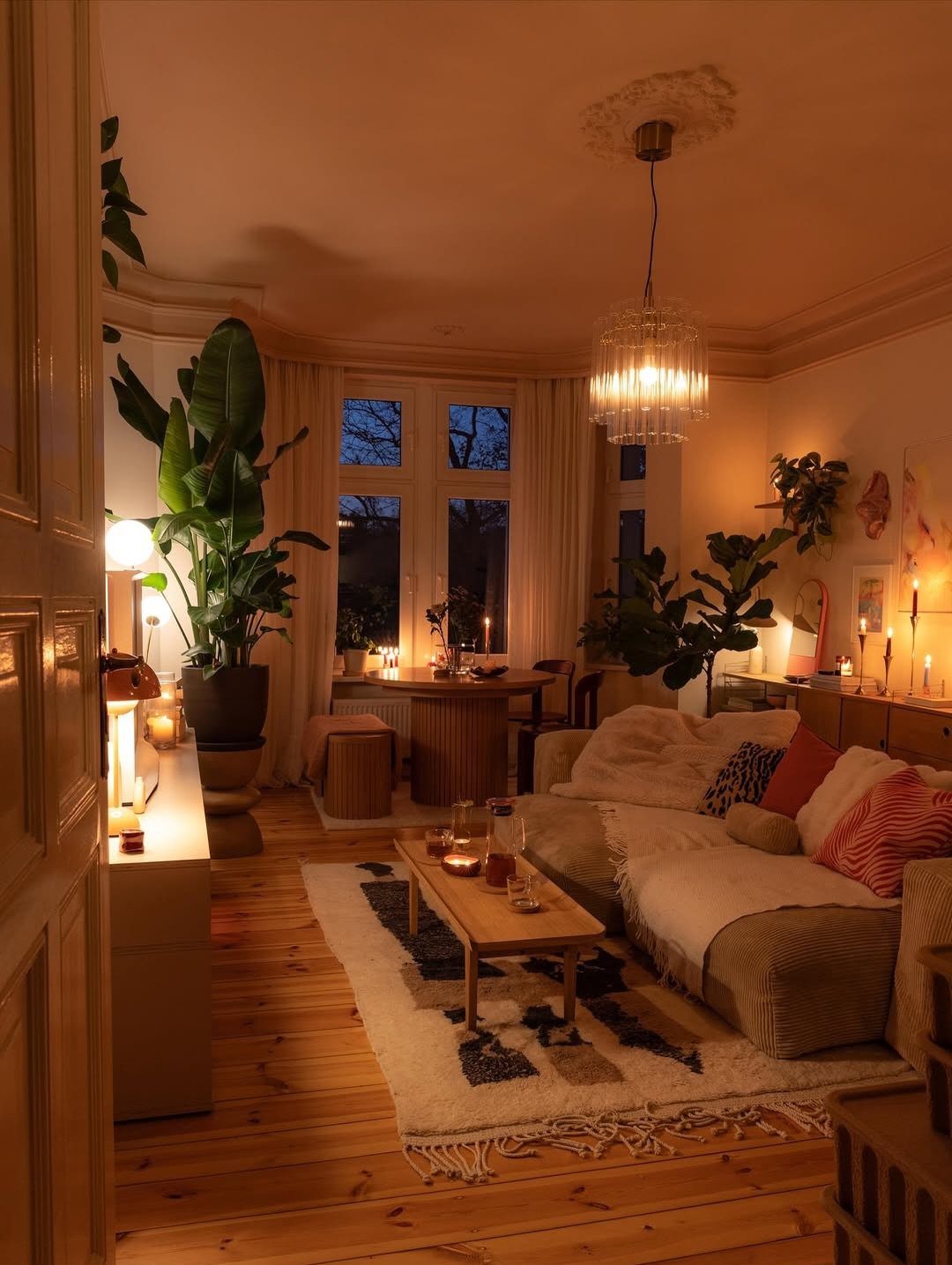A cozy living room with a beige sofa, wooden furniture, indoor plants, and warm ambient lighting.