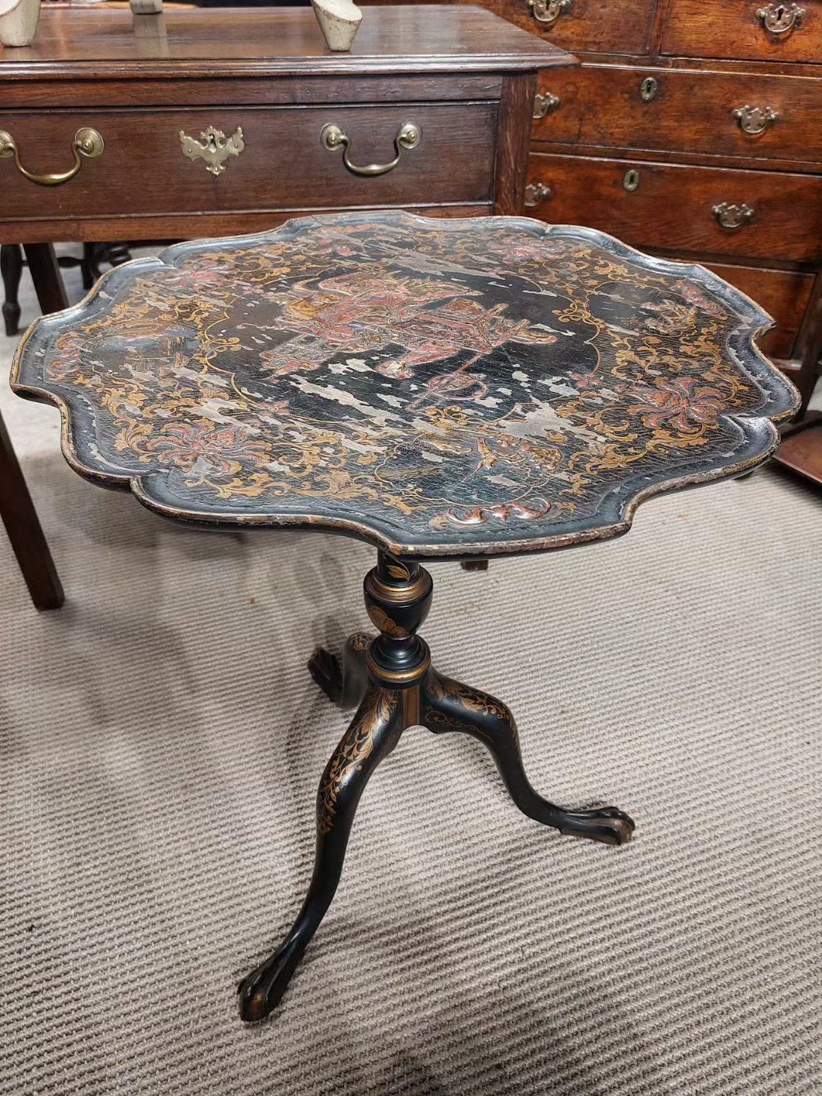 An antique scalloped side table with a dark, patterned decorative top on a carpeted floor.