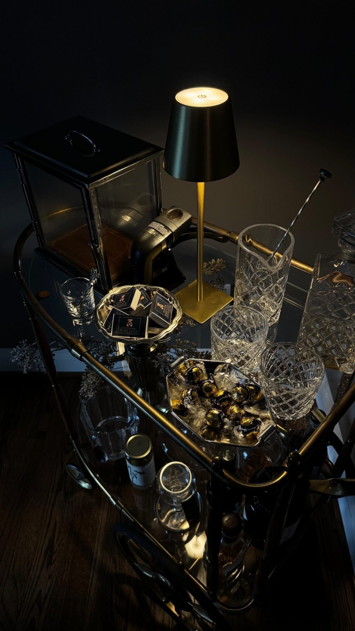 Gold bar cart with crystal glassware, a small lamp, and a black lantern on glass shelves.