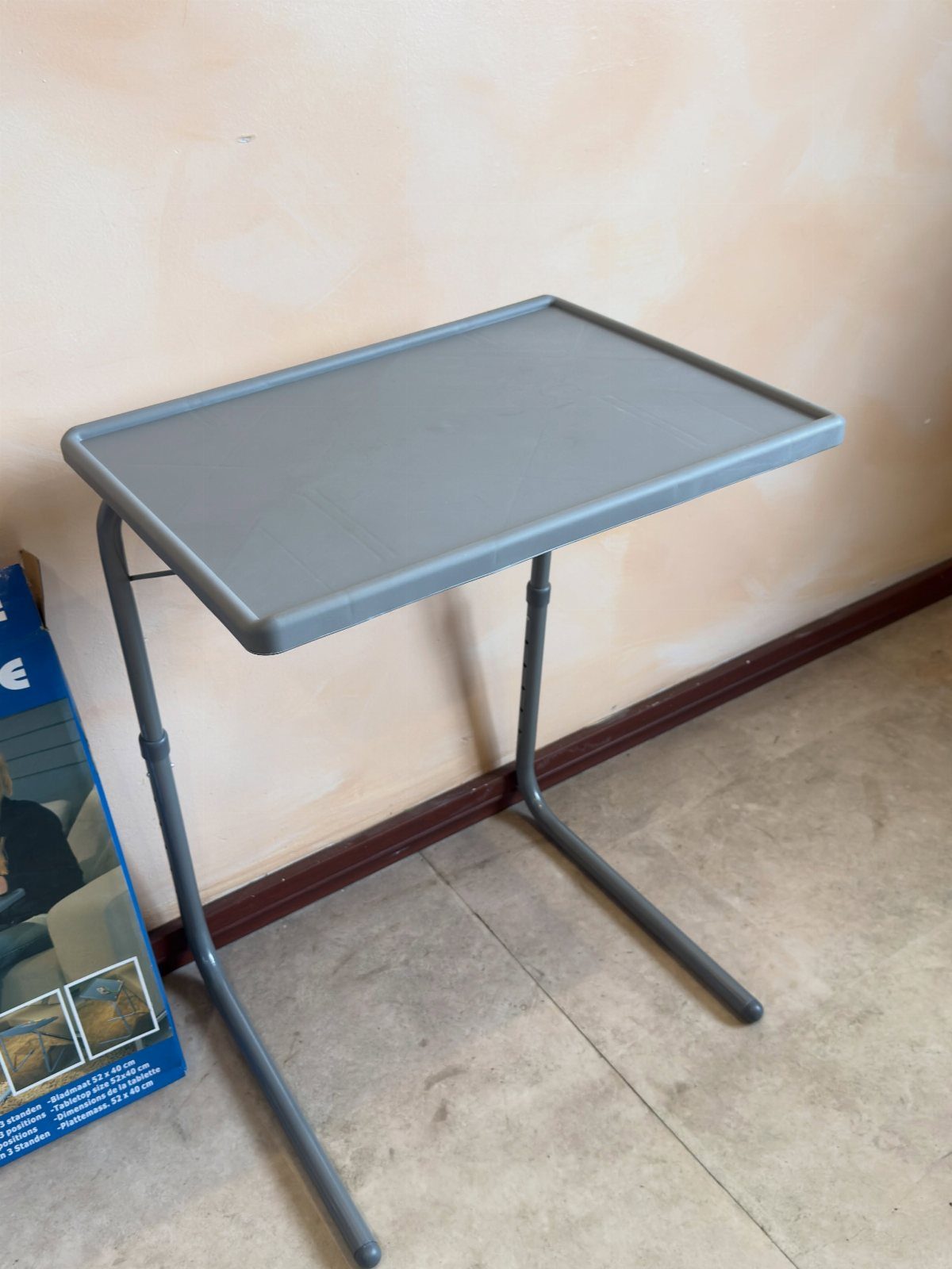 A grey plastic foldable tray table standing against a beige wall on a tiled floor.