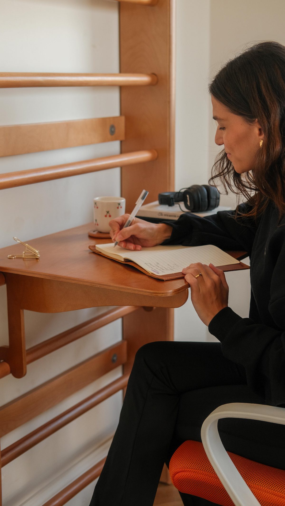 Woman writing in a notebook at a wooden ladder-style desk mounted on a wall.
