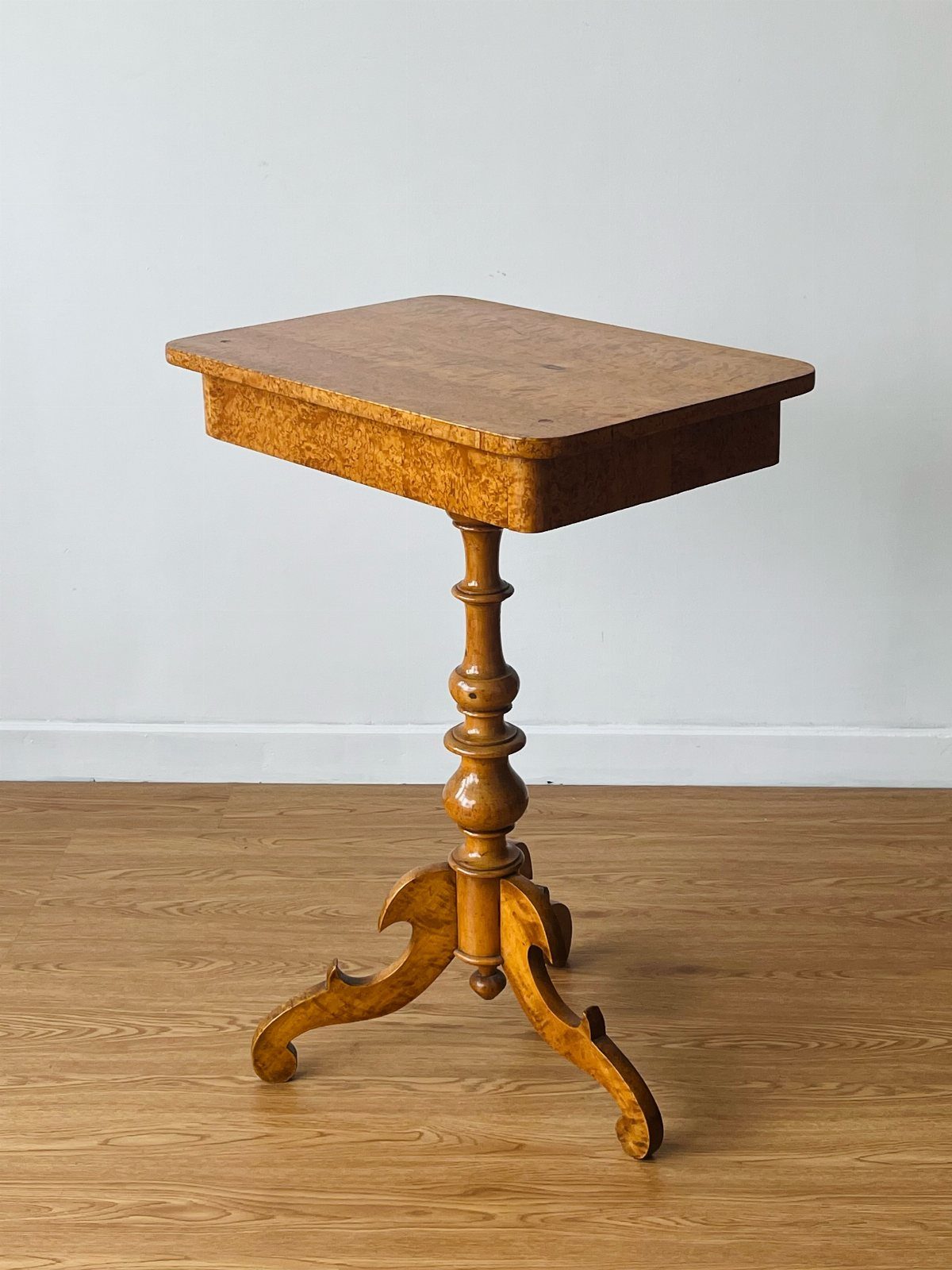 A wooden pedestal table with a square top and decorative tripod base on wood flooring.
