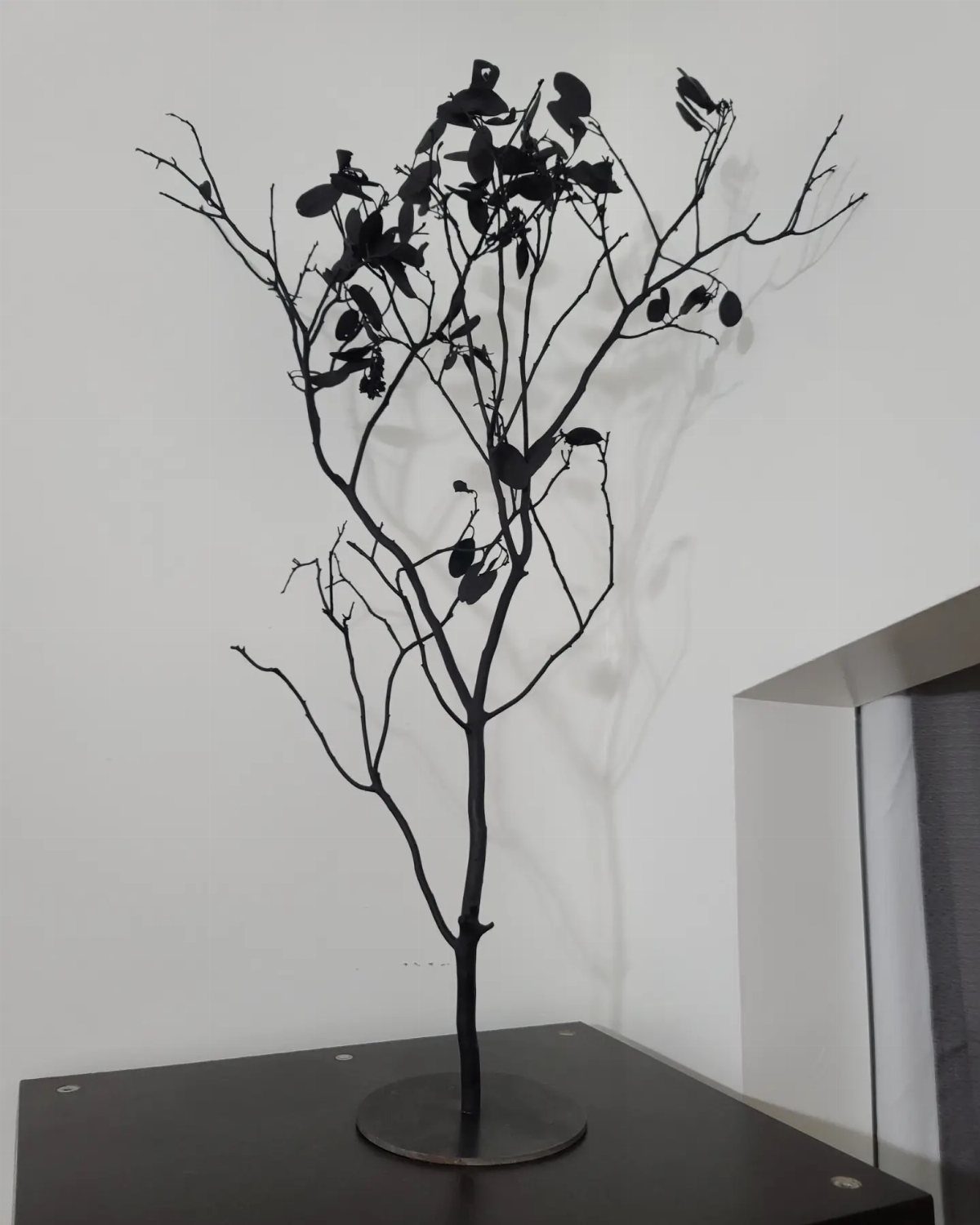 Black decorative branch sculpture with dark accents standing on a dark table against a white wall.