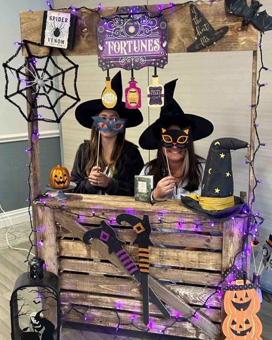 Two women wearing witch hats and masks pose behind a decorated Halloween themed photo booth.