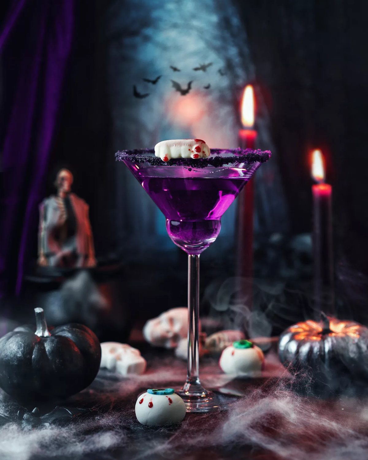 A purple Halloween cocktail garnished with an eyeball, surrounded by black pumpkins and candles.
