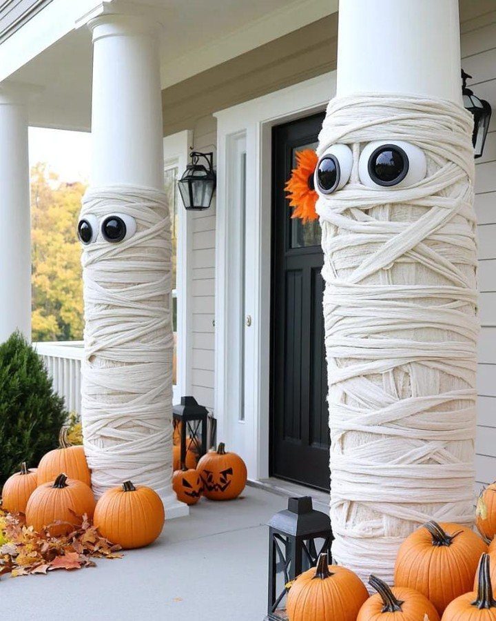 Mummy-wrapped porch columns with large eyes surrounded by orange pumpkins.