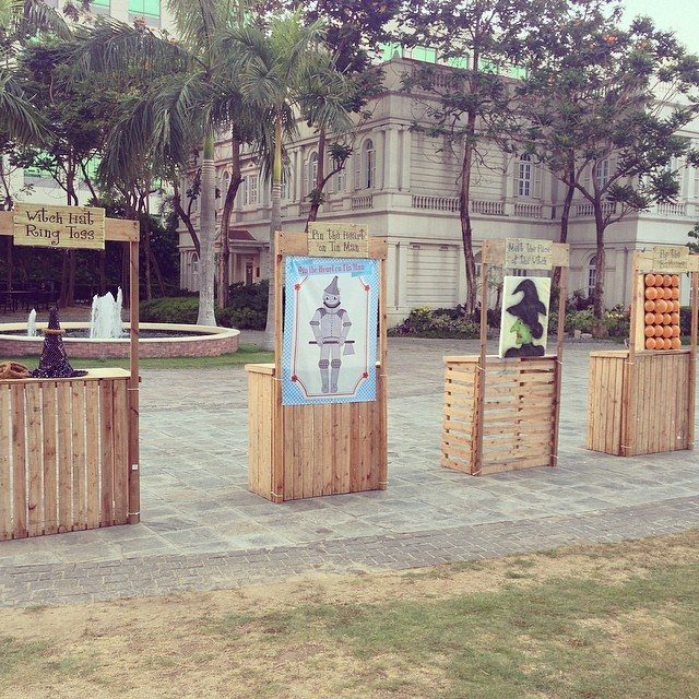 Wooden carnival game booths arranged on a stone patio with palm trees in the background.