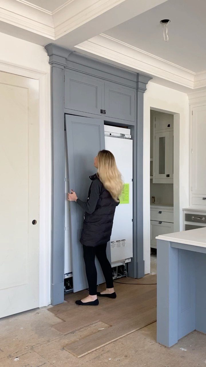 A woman opening a grey cabinet panel to reveal a white refrigerator hidden behind it.