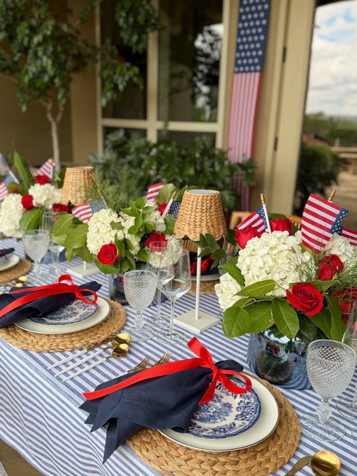 Patriotic table setting with white hydrangeas, red roses, small flags, and blue striped linens.