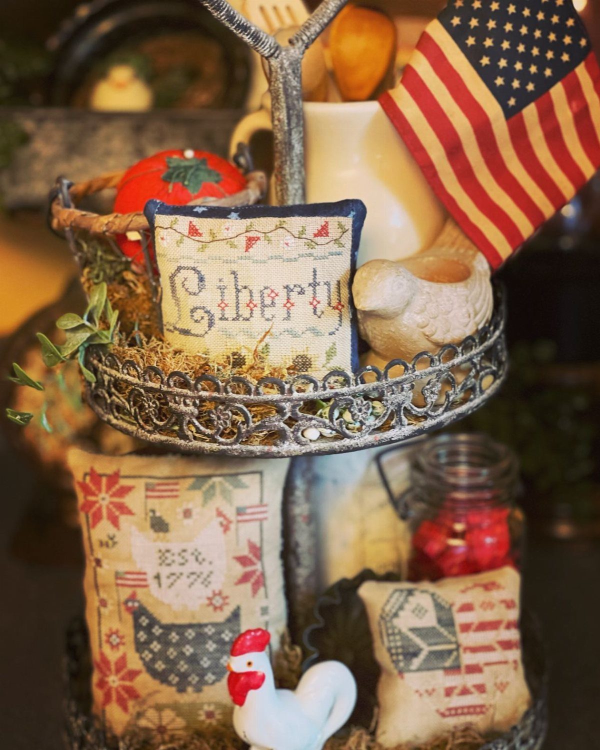 A metal tiered tray decorated with an American flag, Liberty pillow, and rooster figurines.