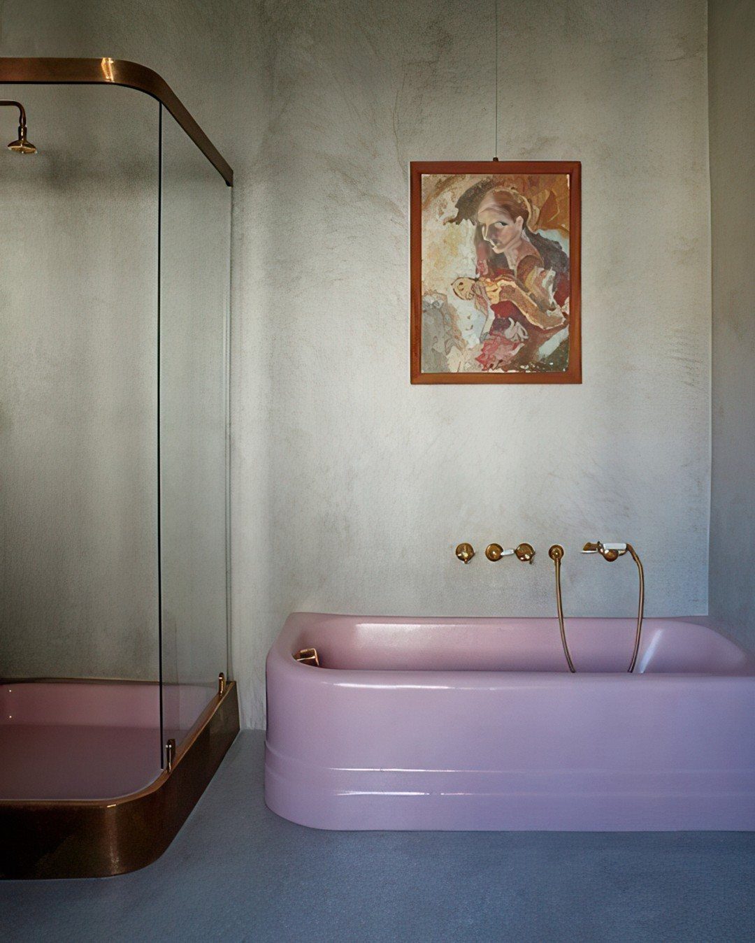 A pink freestanding bathtub with gold fixtures positioned below a framed painting in a grey bathroom.