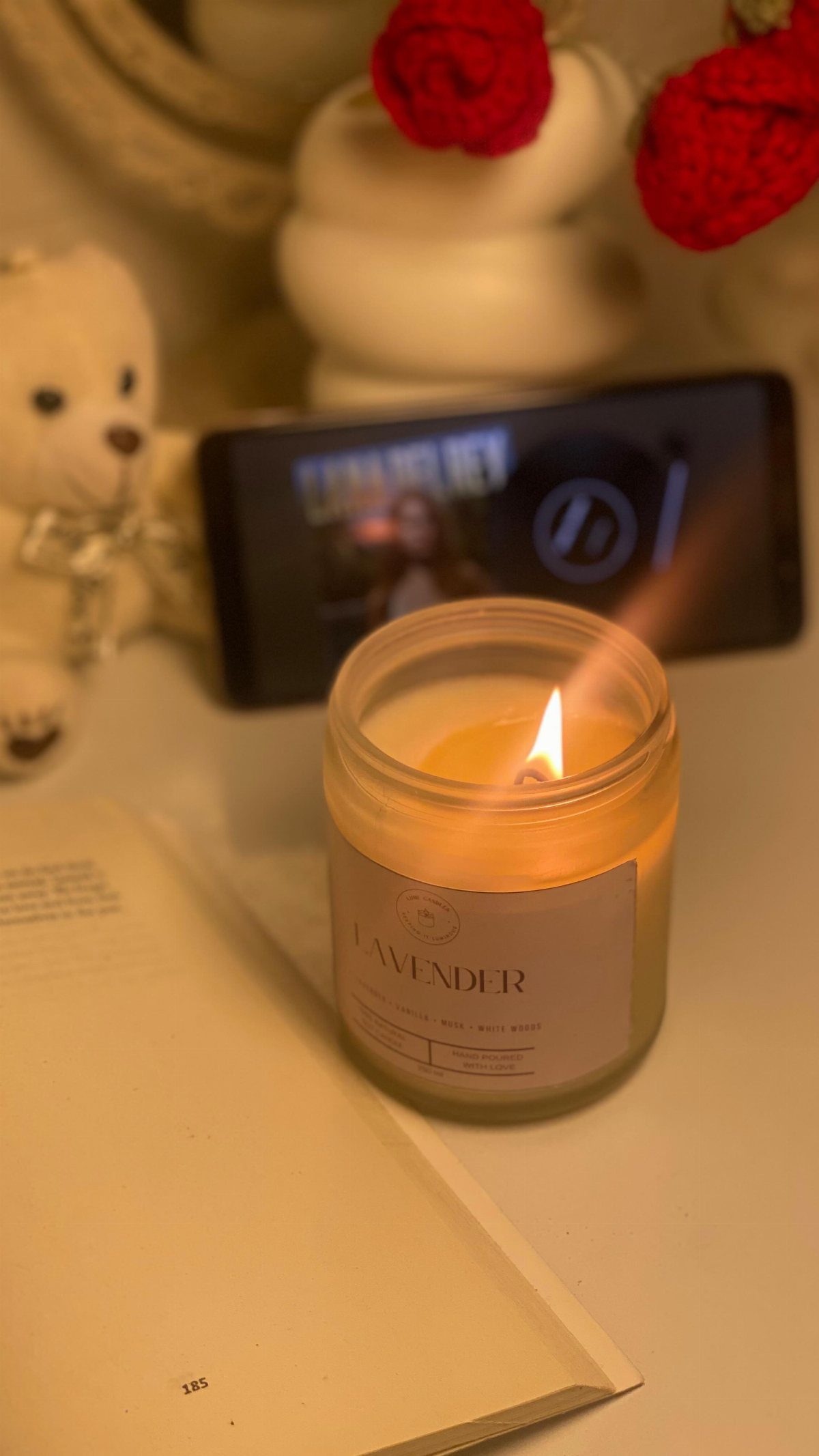 A lit lavender scented candle glowing next to an open book on a white table.