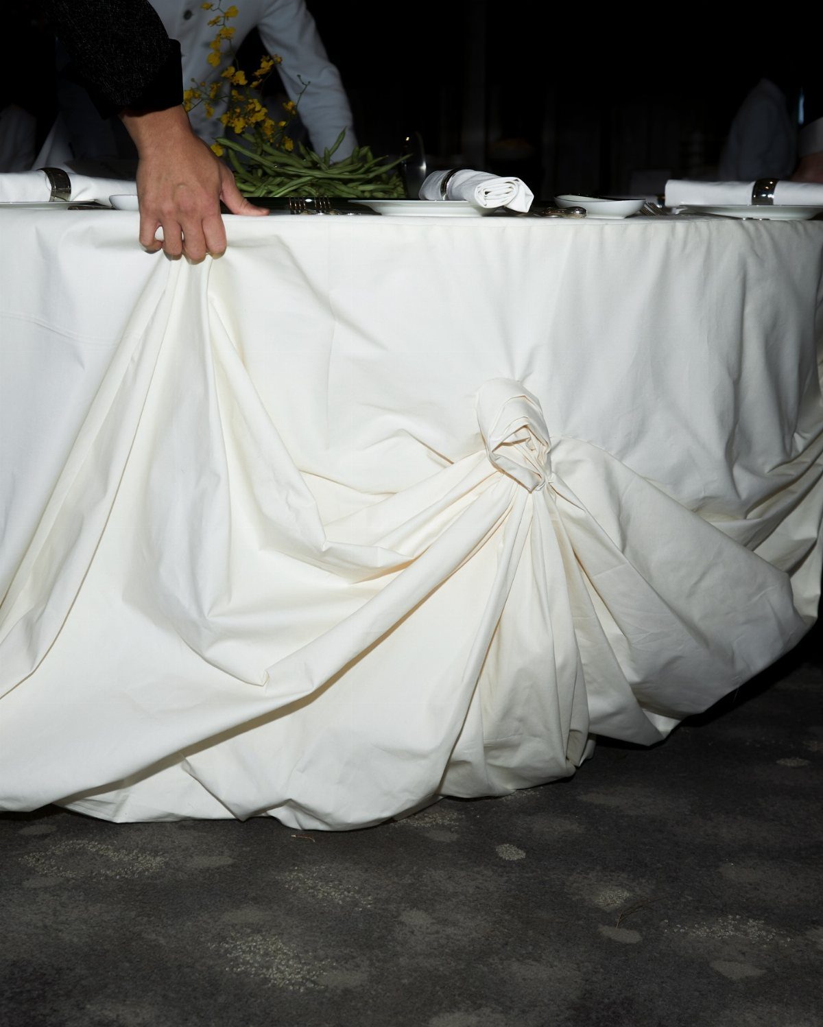 A hand adjusting a white tablecloth gathered into a decorative knot on a long table.