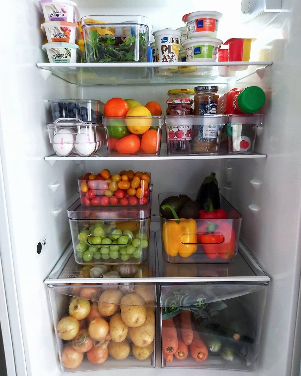 Open white refrigerator filled with organized food containers, fresh fruits, vegetables, and clear plastic storage bins.
