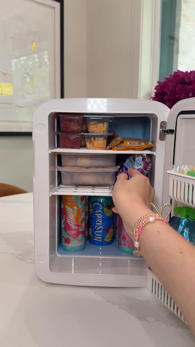 Open white mini fridge filled with snacks and colorful drinks on a white counter.
