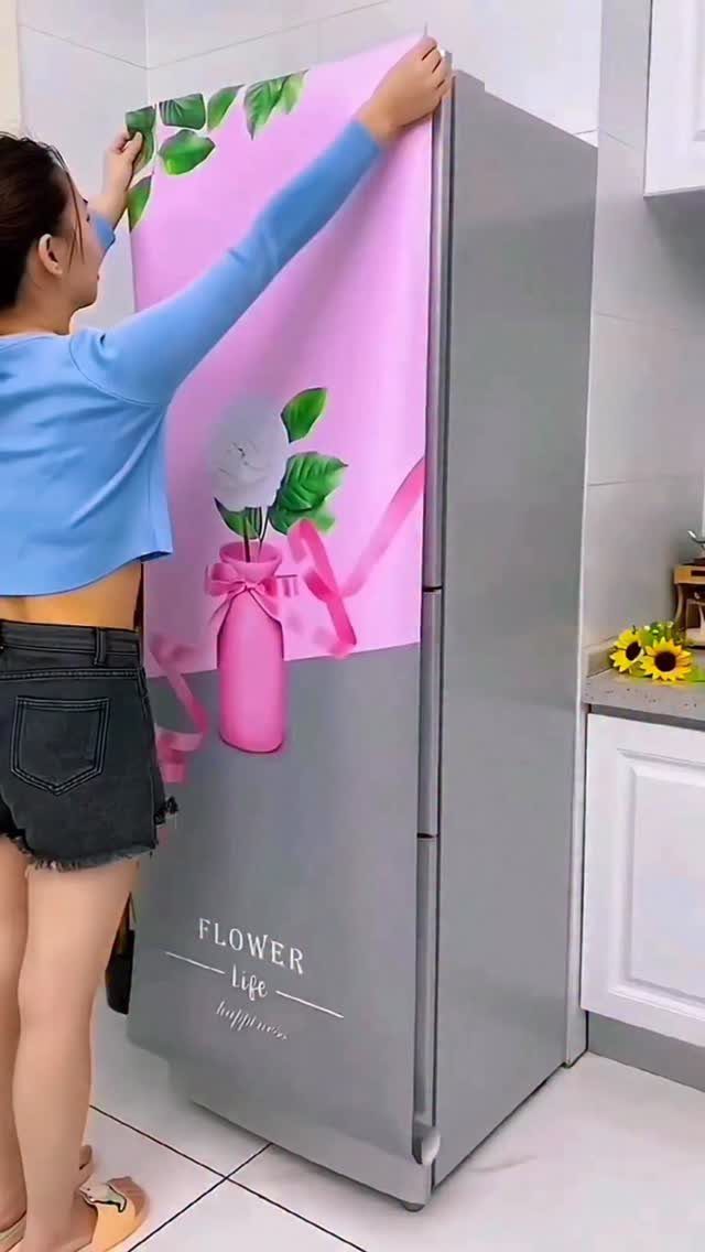 A person applying a pink floral vinyl wrap to a grey refrigerator in a kitchen.