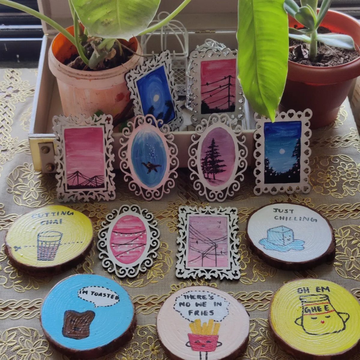 Ornate white miniature frames and painted wooden discs arranged on a gold patterned surface near plants.