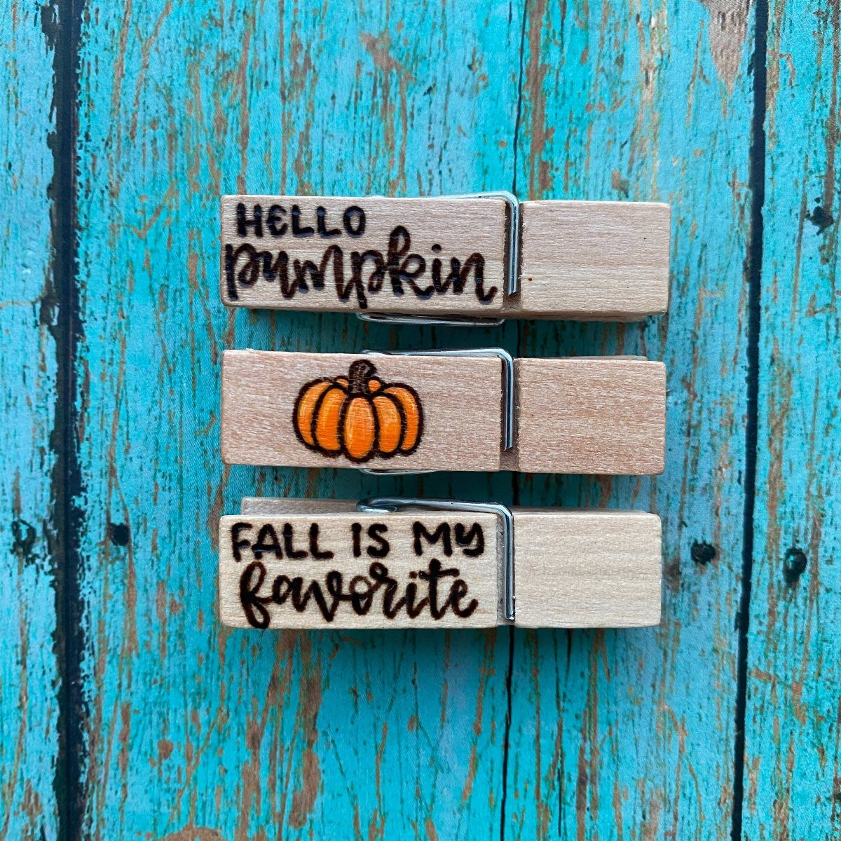 Three decorated wooden clothespins with pumpkin and fall text on blue wood.