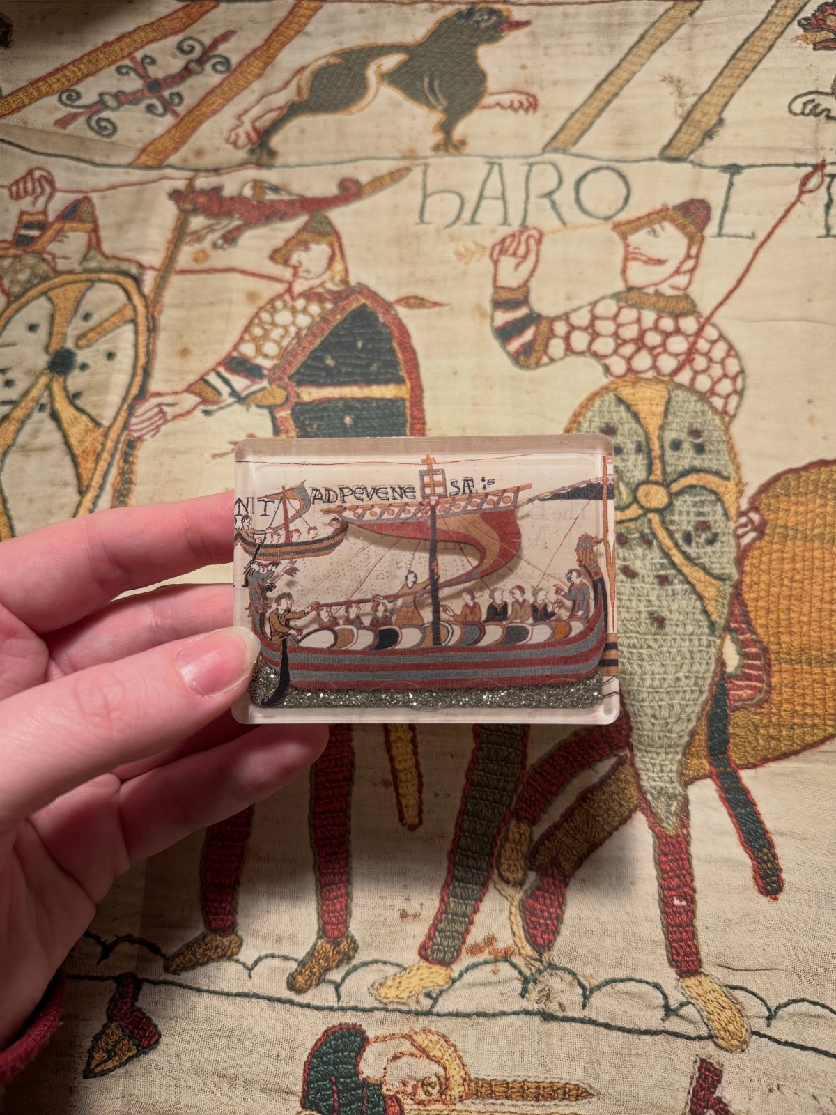 A hand holding a square Viking ship magnet decorated with red and gold glitter.