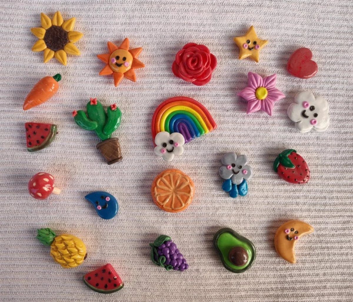 Colorful handmade clay magnets in various shapes like rainbows, fruits, and flowers on white fabric.
